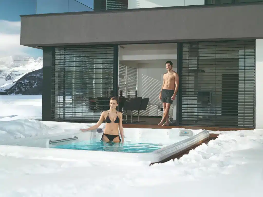 A swimmer stands in an inground swim spa surrounded by snow while another swimmer looks on from the adjacent patio. With proper care, the answer to " Can You Use a Swim Spa in Winter?" is "Yes."