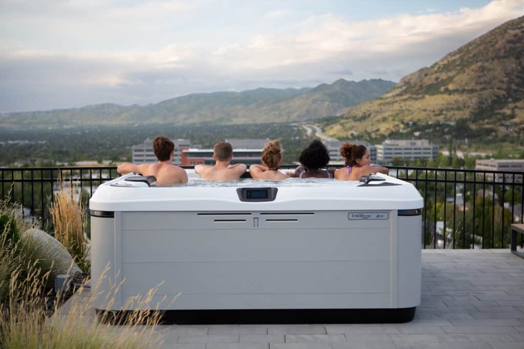 A group of five friends overlooks a city as they relax in a gray hot tub. Hot tub capacity is an important thing to consider when buying a hot tub.